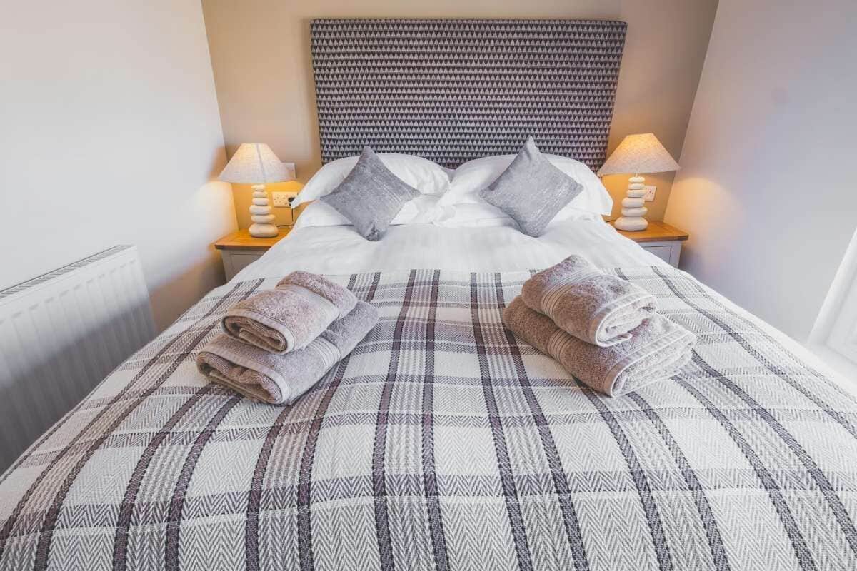 A neatly made bed with a plaid bedspread, two pillows, and two rolled towels on the side.