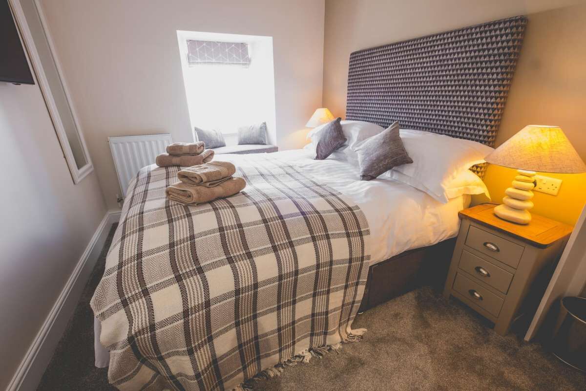A neatly made bed with a plaid blanket, a bedside lamp, and a stack of folded towels.