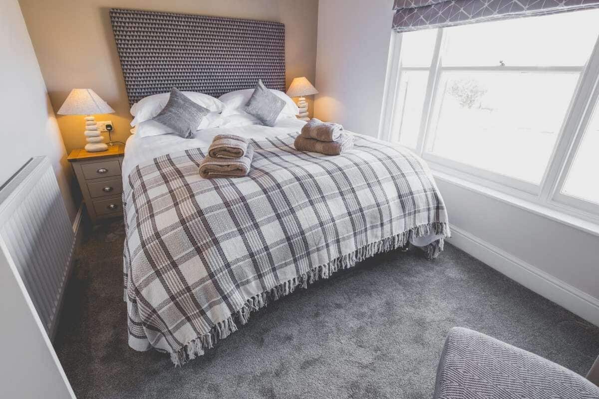A neatly made bed with a plaid blanket, two pillows, and folded towels on a carpeted floor.