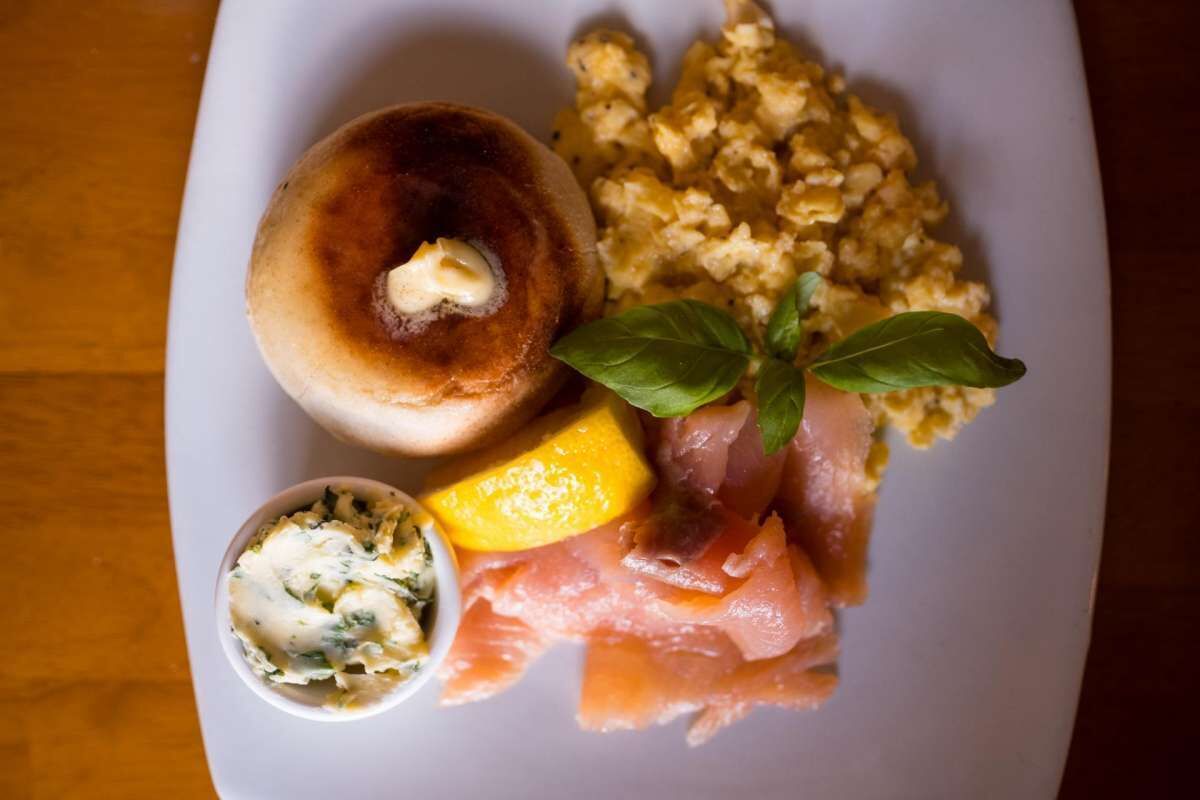 Un plato con un bagel, huevos revueltos, salmón ahumado, un cuenco pequeño de queso crema y una hoja de albahaca.