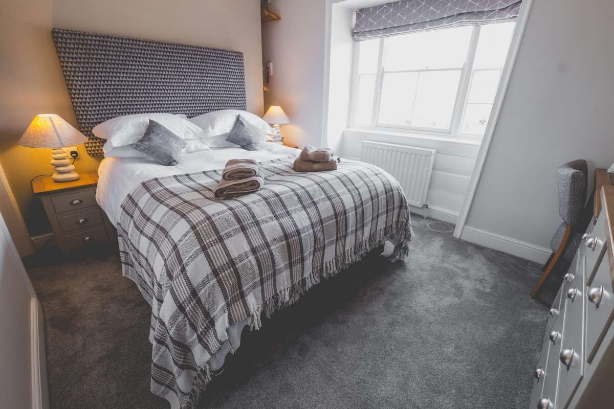 A bedroom with a bed, two bedside lamps, a radiator, a window with a valance, and a dresser.
