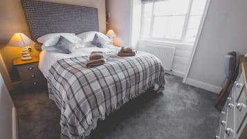 A bedroom with a bed, two bedside lamps, a radiator, a window with a valance, and a dresser.