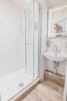 A modern bathroom with white subway tiles, a glass shower enclosure, a wooden floor, and a white sink with a mirror above it.