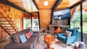Ein gemütliches Wohnzimmer mit Kamin aus Stein, einem an der Wand befestigten Fernseher, einem blauen Sessel und einem Holztisch.