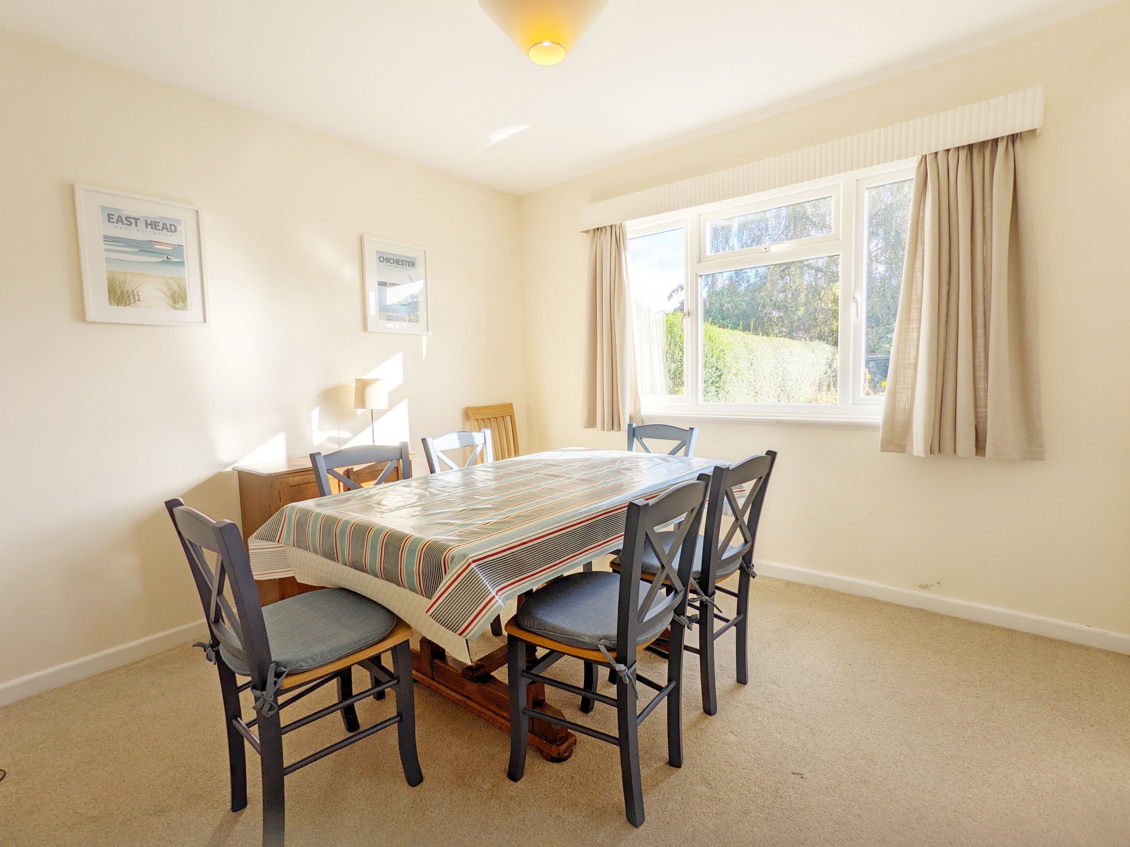 A dining room with a table set for four, surrounded by chairs. There are two framed pictures on the wall, a window with curtains, and a carpeted floor.