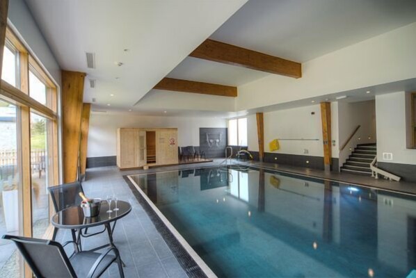 En indendørs swimmingpool med glasvæg, et bord med stole og en trappe.