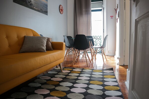 Uma sala de estar com um sofá amarelo, um tapete estampado e um espaço de jantar com cadeiras e uma mesa.