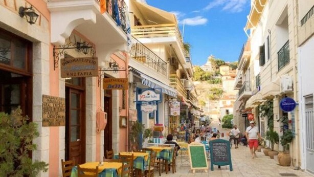 Une rue étroite bordée de terrasses, de boutiques et de panneaux, le tout dans un cadre méditerranéen.