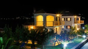 Un bâtiment en pierre de deux étages, doté de balcons et d’un jardin bien éclairé la nuit.