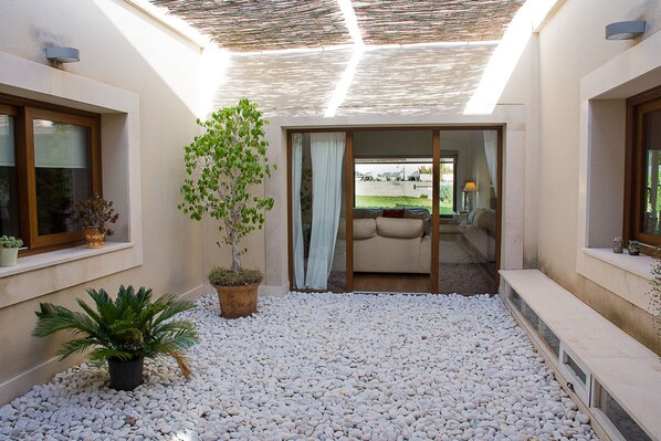 Un interno moderno con zona patio caratterizzata da ciottoli bianchi, una pianta in vaso e una porta scorrevole in vetro che dà su una zona relax con divano e lampada.