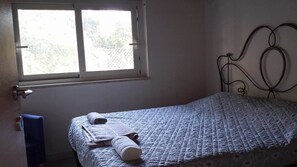 Ein kleines, ordentlich bezogenes Bett mit weißer Decke und zwei aufgerollten Handtüchern. Ein Fenster mit Blick auf Bäume ist zu sehen.