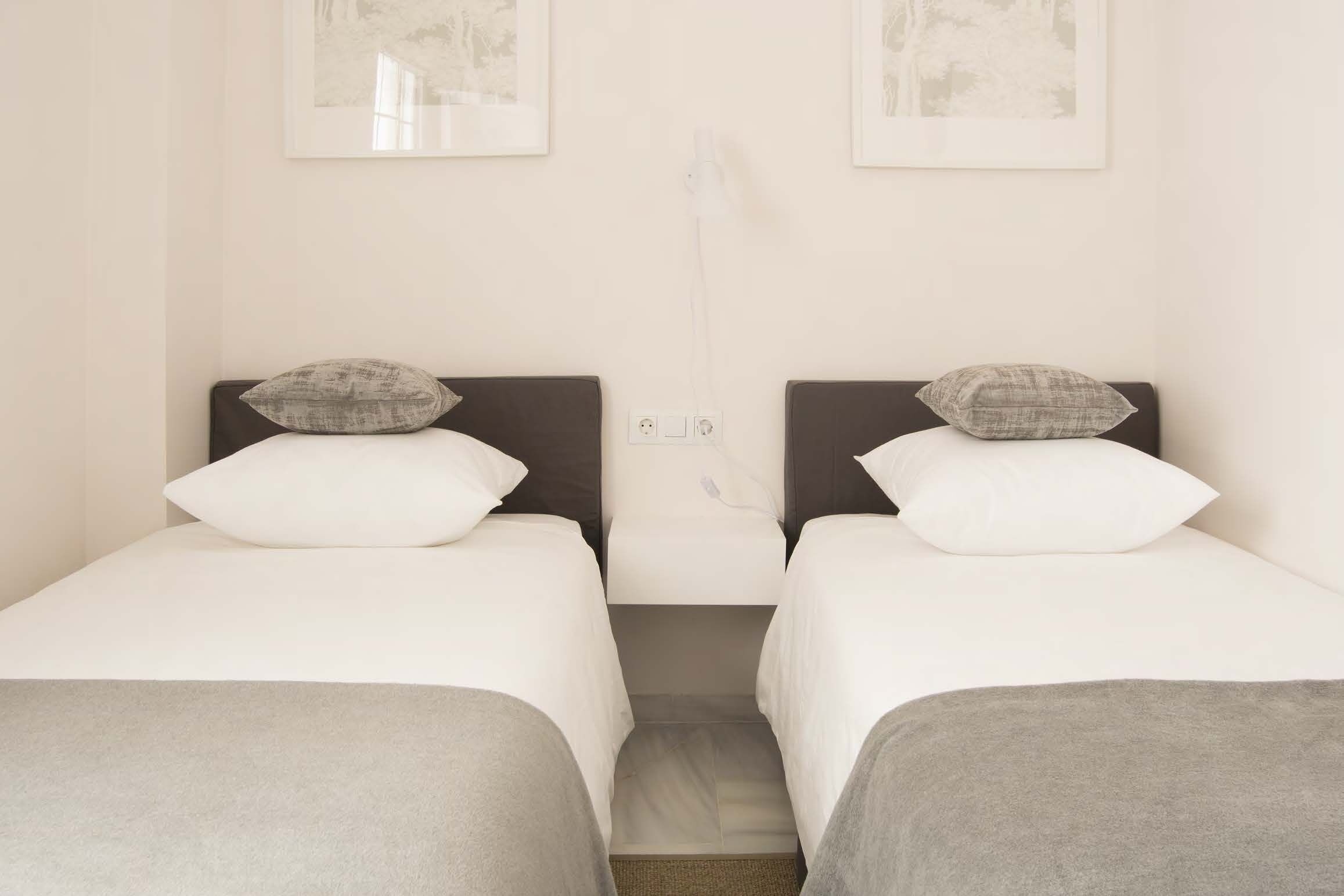 Quarto de hotel com duas camas individuais, cada uma com travesseiro e cobertor. Há duas imagens emolduradas na parede acima das camas.