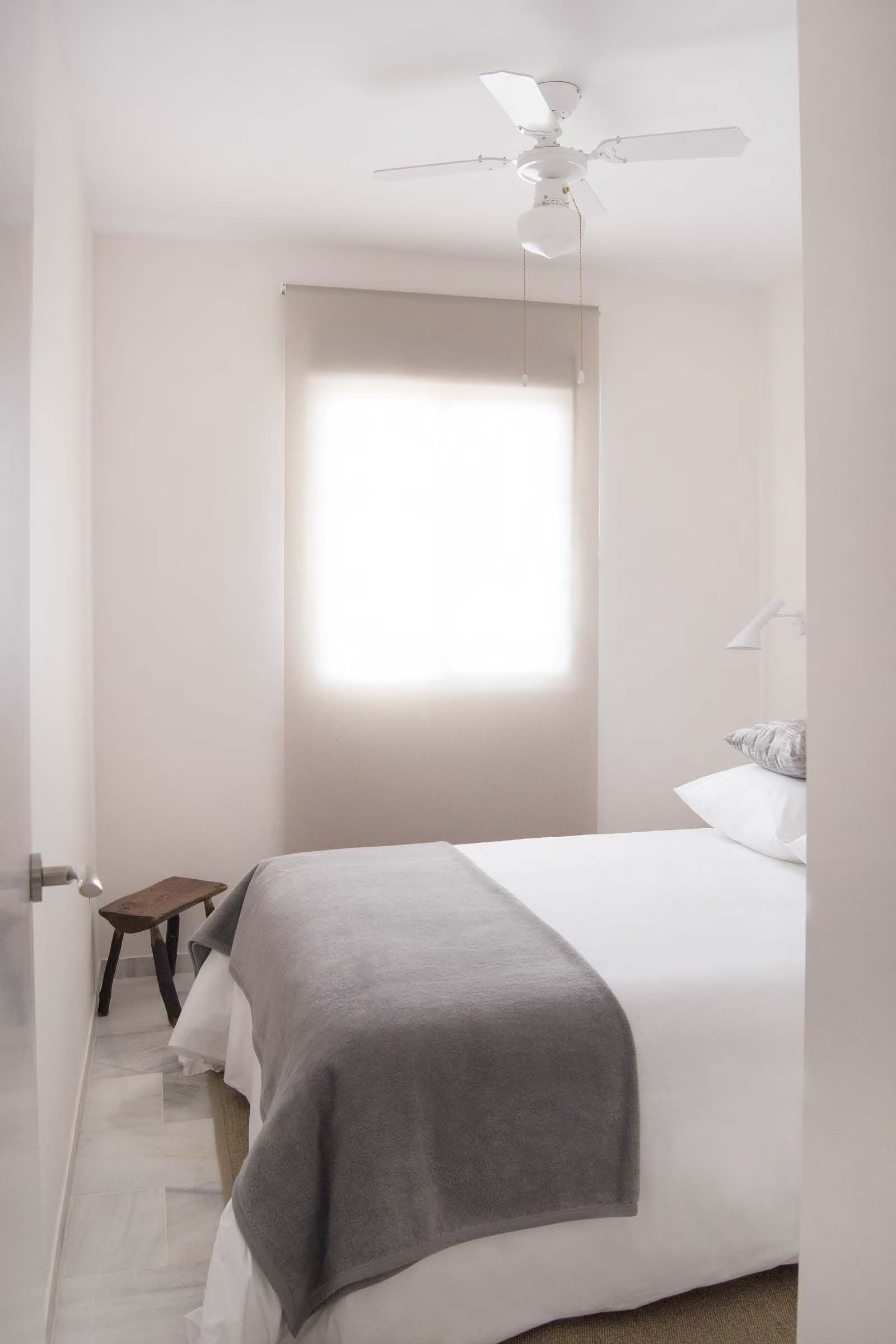 Ein Schlafzimmer mit einem Bett, einem Deckenventilator, einem Fenster und einem kleinen Holzschemel.