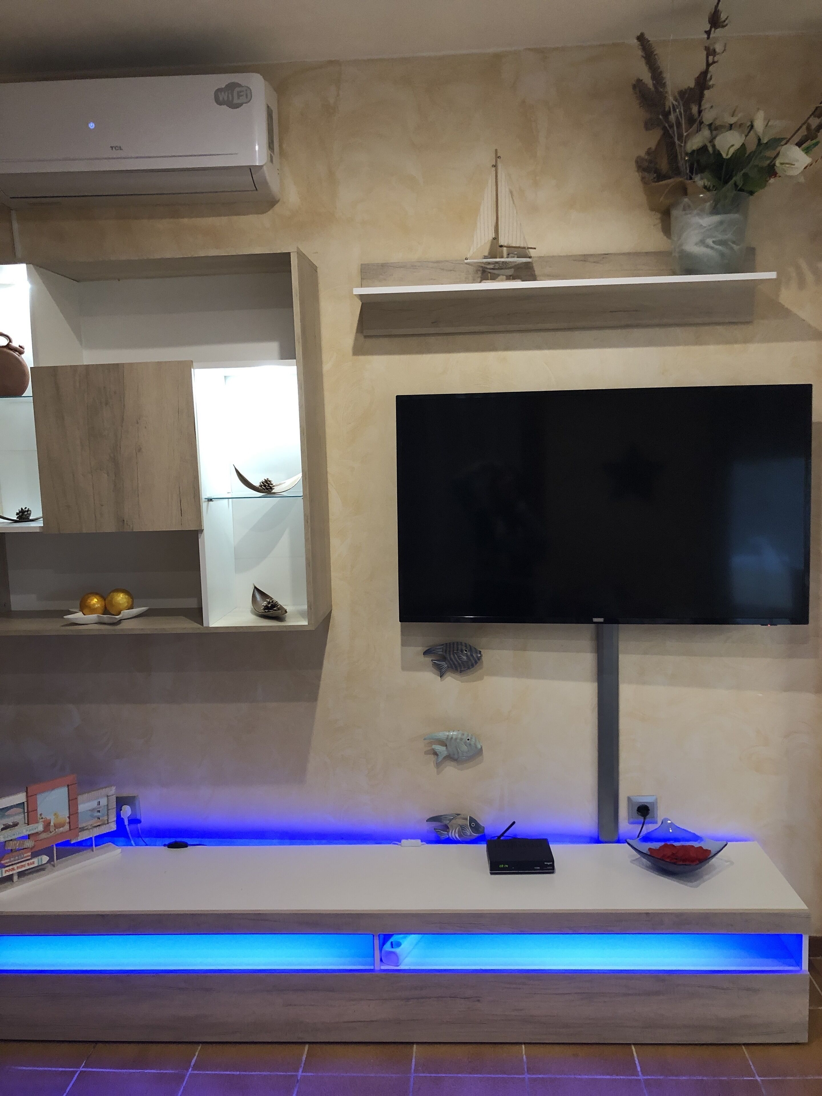 Un salon moderne avec un téléviseur fixé au mur, une étagère en bois ornée d’objets décoratifs et une console intégrée illuminée de lumière bleue.
