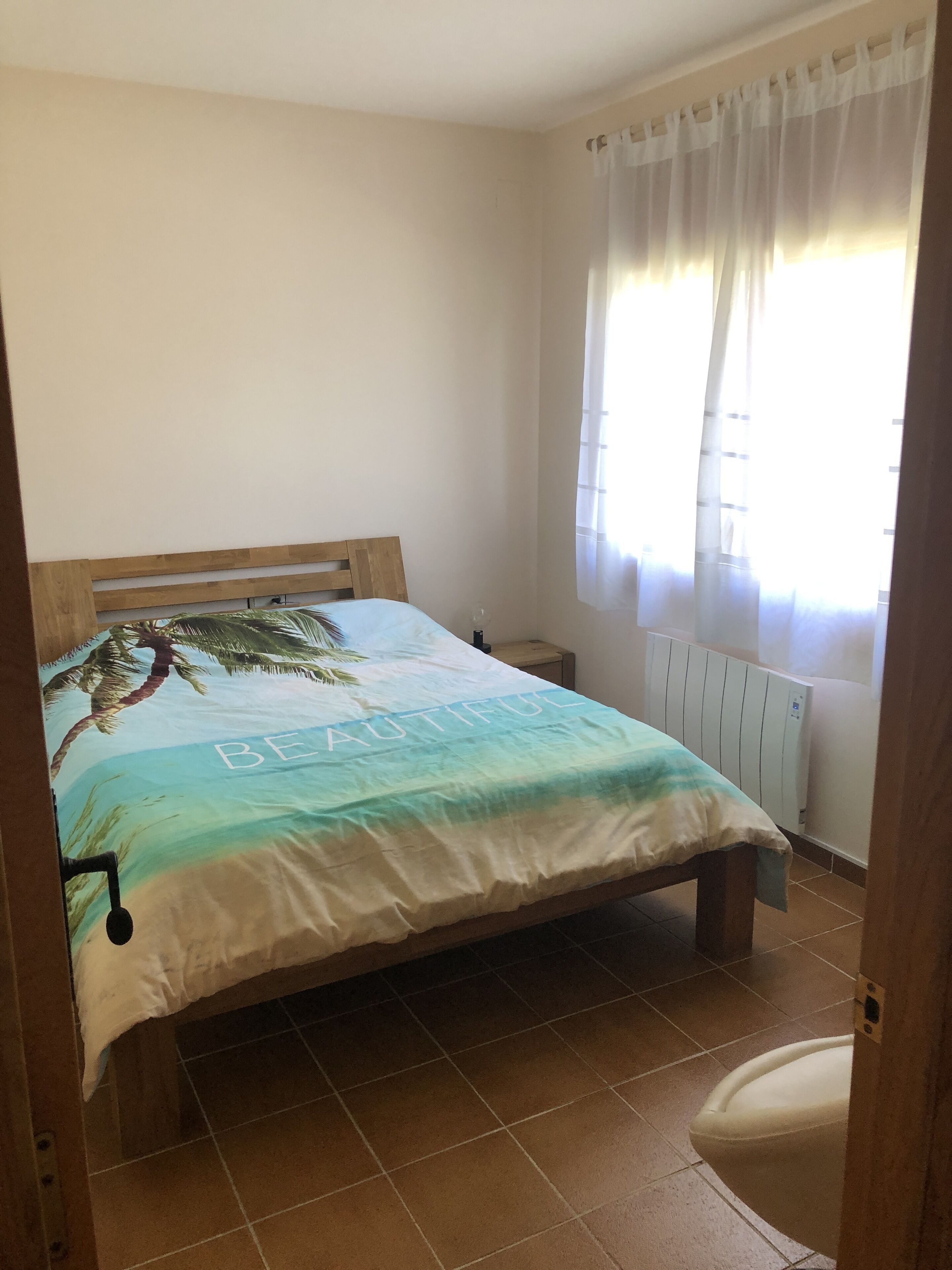 A bedroom with a bed, a nightstand, a window with curtains, and a radiator.