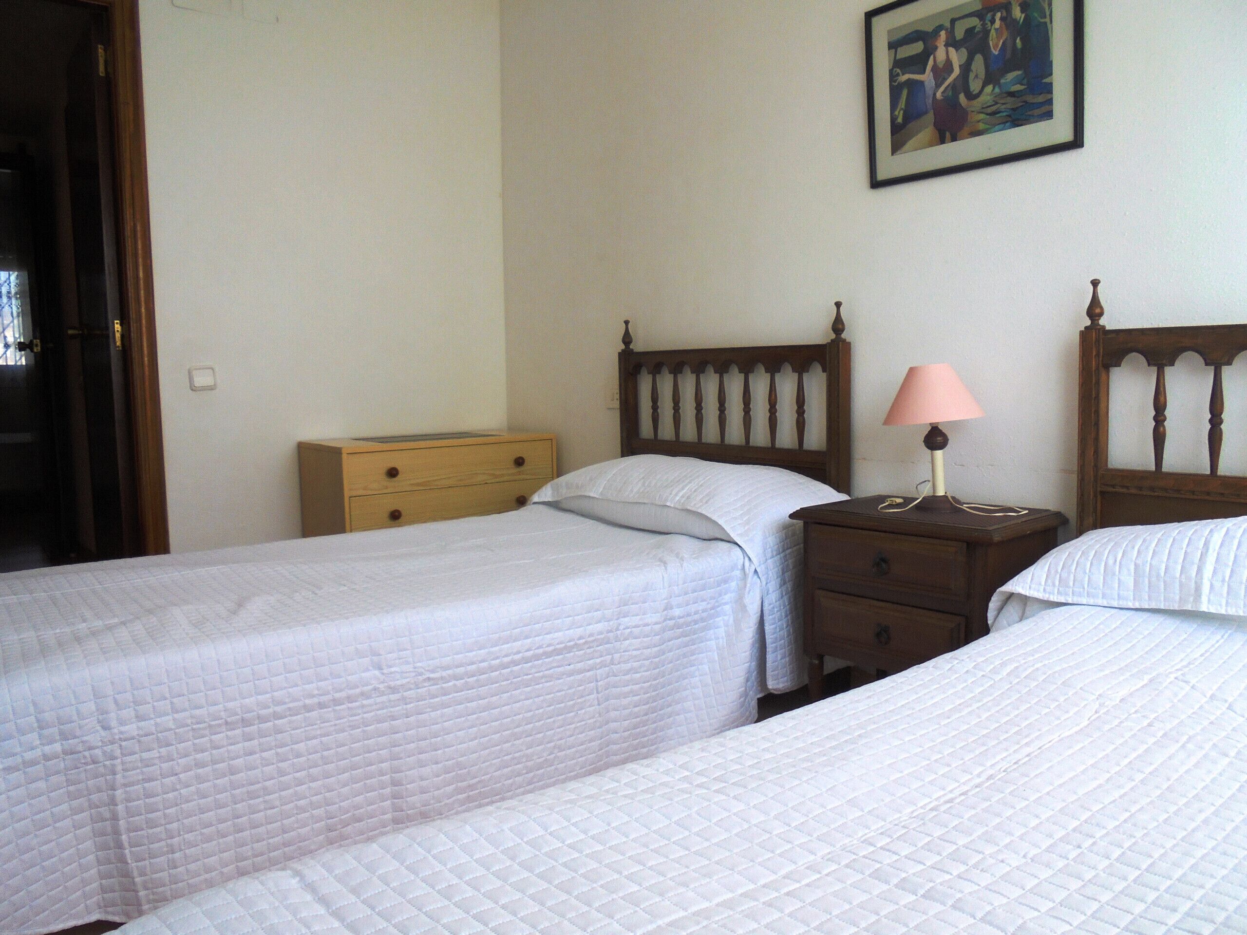 Un dormitorio con dos camas individuales, una mesita de noche de madera con lámpara y un pequeño aparador.