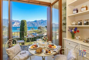 Un balcon avec une table dressée pour le petit-déjeuner, offrant une vue sur un paysage montagneux et une étendue d’eau.