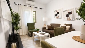 A modern living room with a sofa, armchairs, a coffee table, and a dining table. There are two wall-mounted air conditioning units, a potted plant, and a lamp.