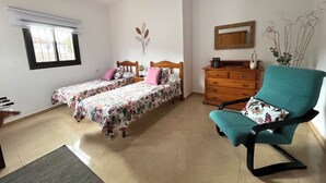 A bedroom with two single beds, a wooden dresser, a teal armchair, and a window with blinds.