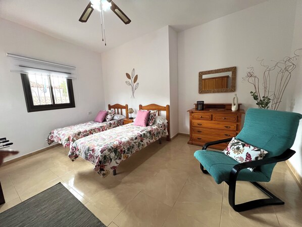 A bedroom with two single beds, a wooden dresser, a teal armchair, and a window with blinds. - Casa Victoria Sunny Terrace & Teide Views (Adeje)