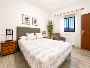 A bedroom with a bed, a nightstand, a chair, a window with curtains, and two wall-mounted pictures.