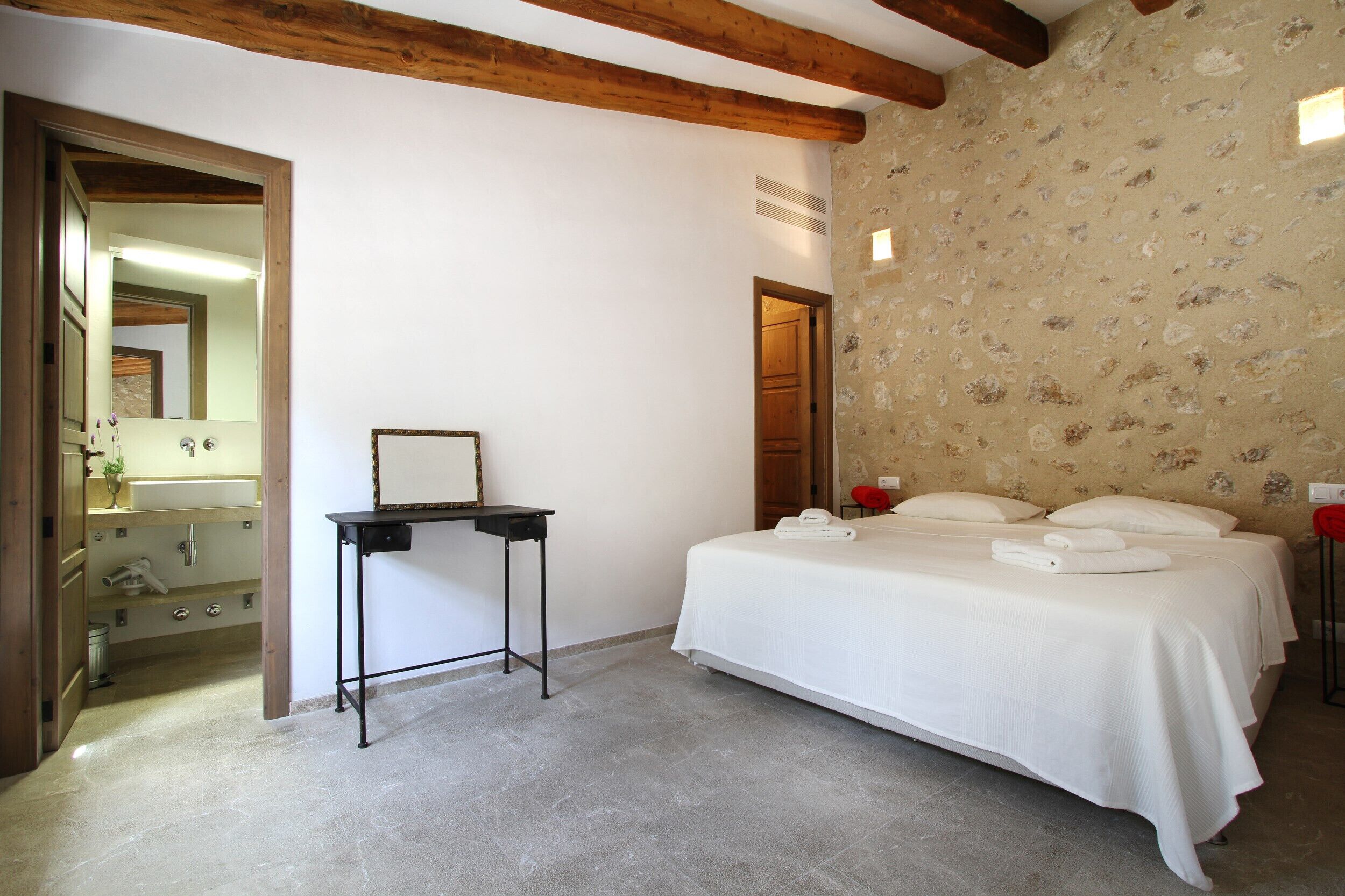 A bedroom with a bed, a small table, a mirror, and a bathroom visible through an open door.