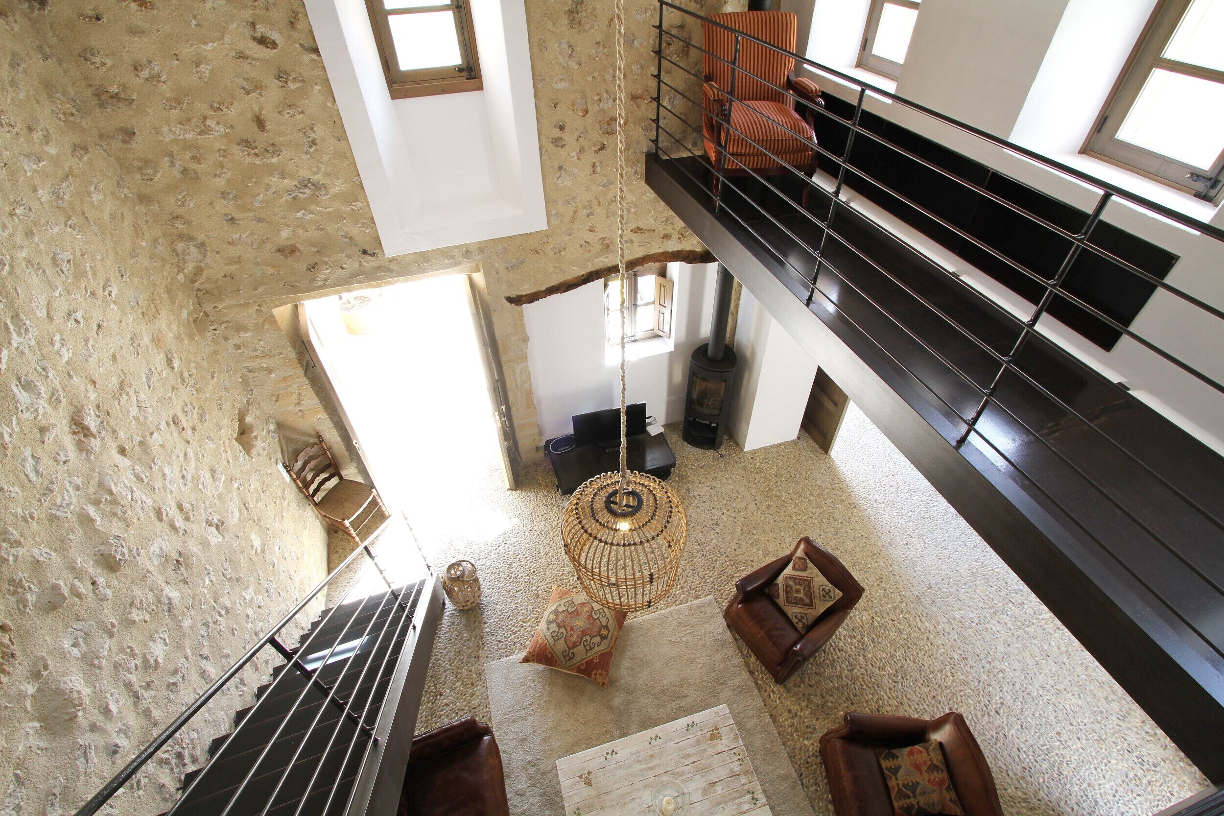 A high-ceiling room with a stone wall, a wooden floor, a hanging light fixture, and a staircase leading to a balcony with chairs.