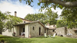Ein Haus aus Stein mit Ziegeldach, einer kleinen Veranda und einem Rasenstück mit zwei weißen Liegestühlen.