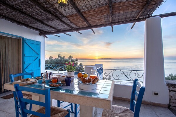 A terrace with a table set for breakfast overlooking the sea at sunset.
