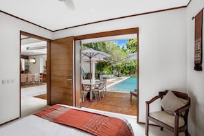 A bedroom with a large bed, a wooden door leading to a patio with a pool, a dining area, and a chair.