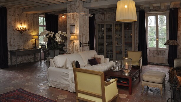 A cozy living room with a white sofa, a wooden chair, a small table with a candle, and a view of the outdoors through a window. - La Demeure de l'Ile (Craon)