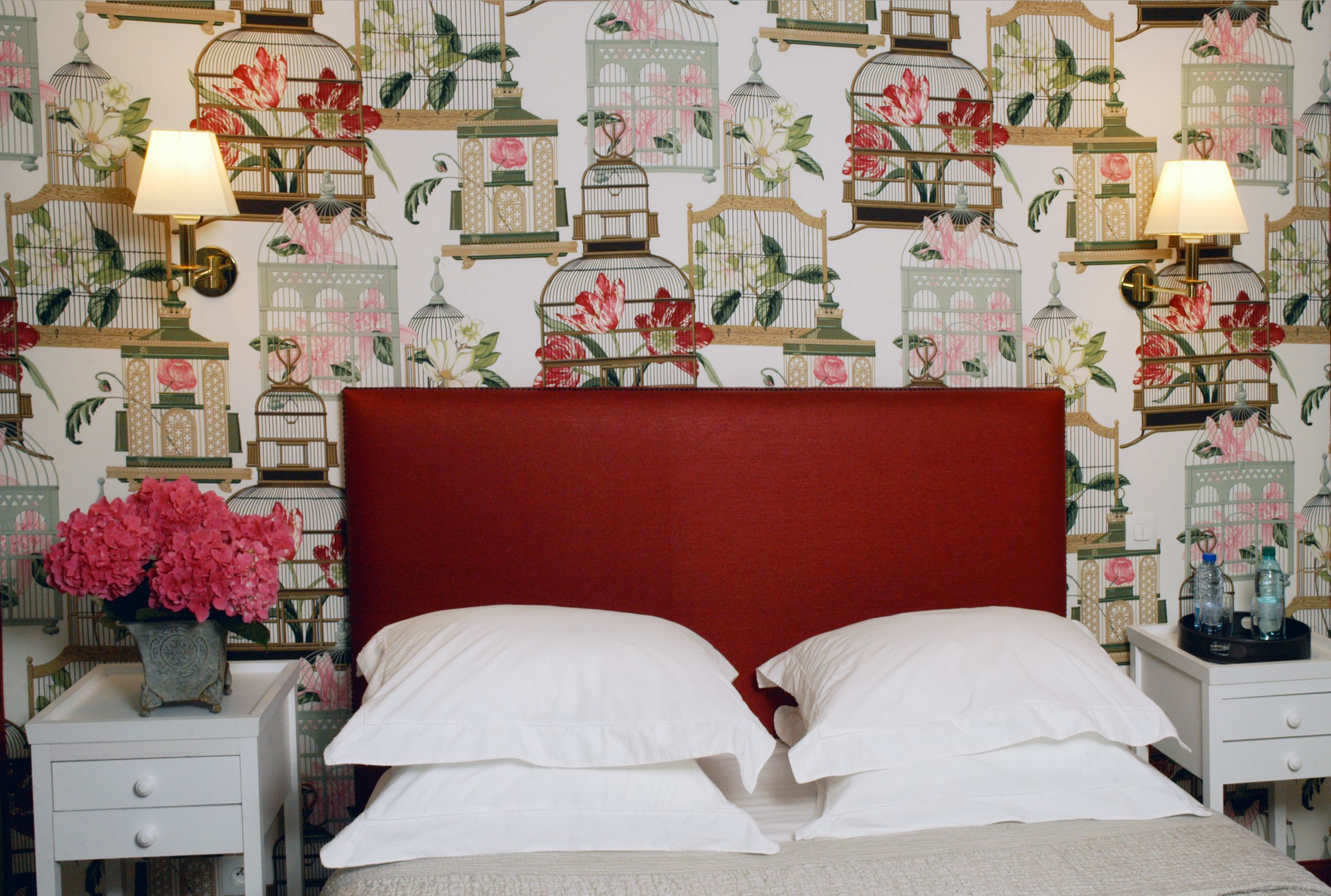 A bedroom with a red headboard, white pillows, and a floral-patterned wallpaper featuring birdcages and flowers. There are two bedside tables with lamps and a vase of flowers on one table.