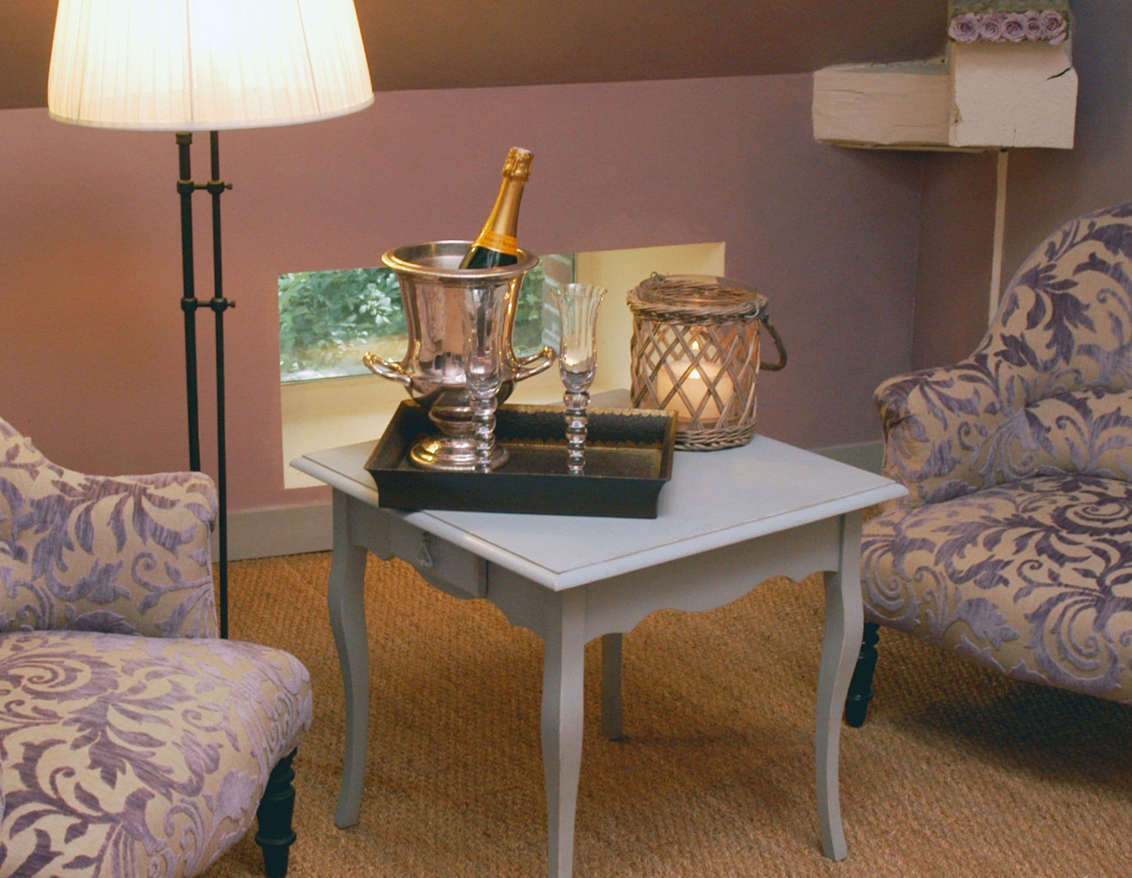 A room with a table set with a bottle of champagne, glasses, and a candle holder. There are two patterned armchairs and a floor lamp.