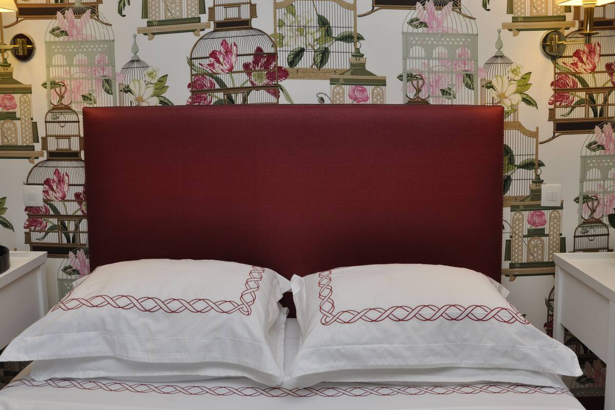A bed with a red headboard and white pillows with red patterns, set against a wall with birdcage wallpaper.