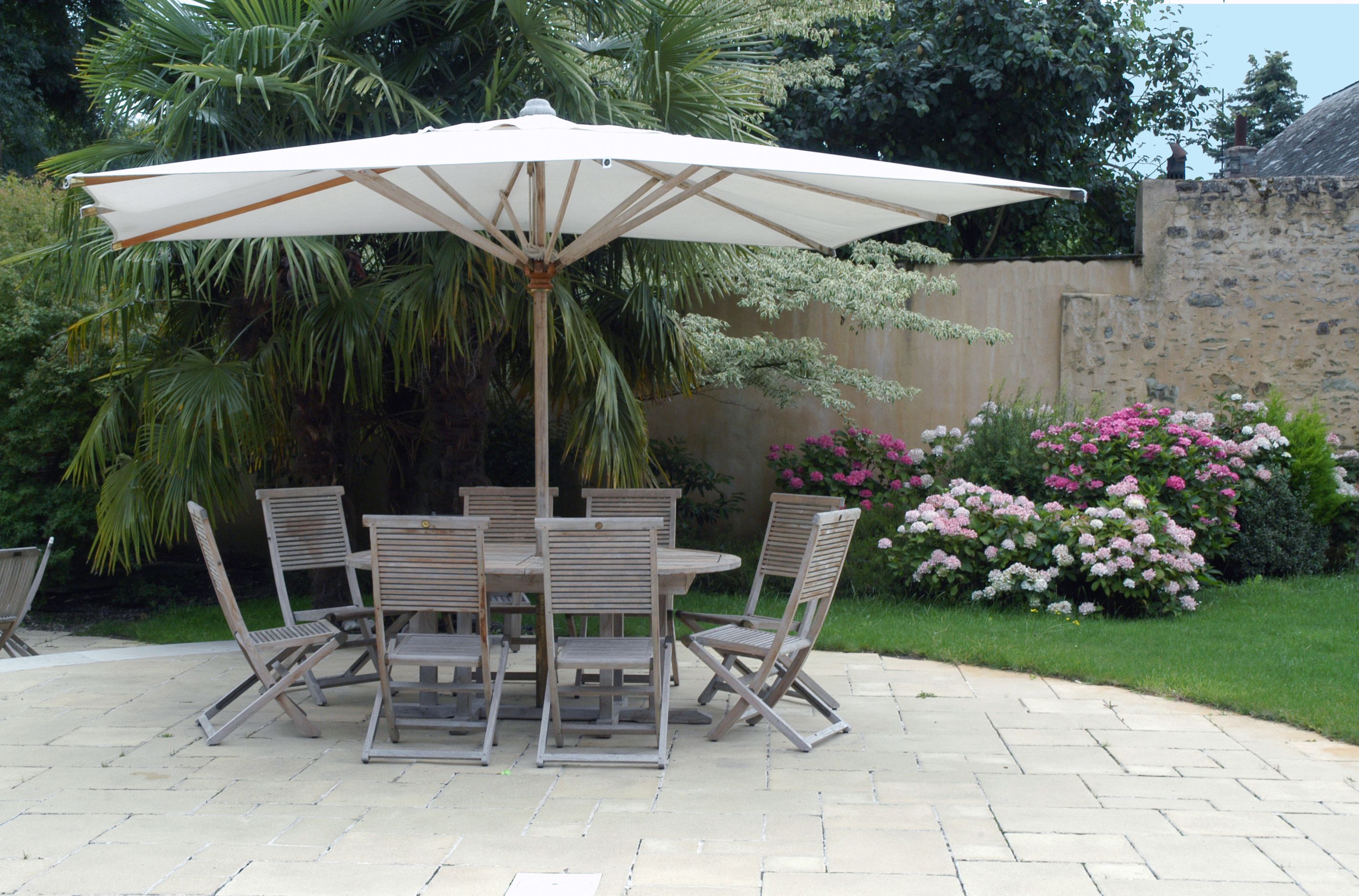 A patio with a table and chairs under a large umbrella, surrounded by lush greenery and flowering plants.