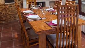 Un comedor rústico con una mesa de madera dispuesta para cuatro personas, rodeada de sillas de madera. Se observa una chimenea de piedra al fondo, y el suelo es de baldosas.