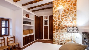 Un dormitorio con pared de piedra, vigas de madera, cama, mesita de noche, lámpara y estanterías empotradas con cestas y un florero.