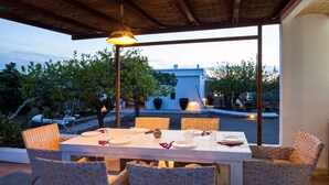 Un espacio de comedor al aire libre con una mesa preparada para cuatro personas, rodeado de sillas de mimbre, bajo una pérgola con una lámpara colgante.