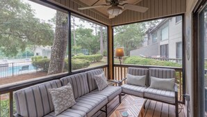 A covered outdoor seating area with a sofa and chairs, a ceiling fan, and a view of a swimming pool and greenery. - 10 Ocean Gate: 2BR, 2.5BA Community Pool, Short Walk to Beach, Free Bikes (Hilton Head Island)