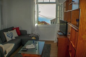 A living room with a sofa, a coffee table, a television, and a view of the sea through a window.