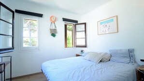 Un dormitorio con una cama, dos ventanas, un reloj de pared, un cuadro enmarcado y una mesita de noche.
