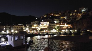 Uma cidade à beira-mar com edifícios iluminados à noite, barcos atracados e uma ponte em primeiro plano.