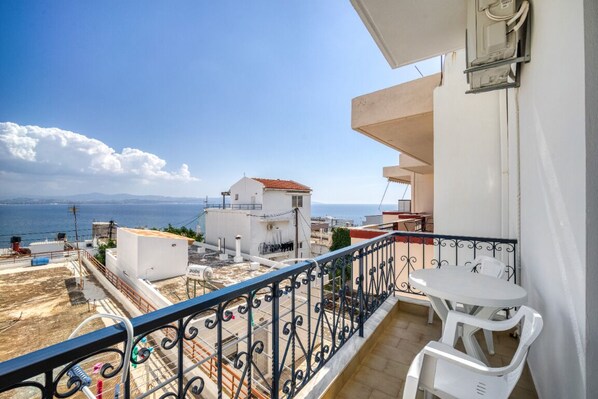 A balcony with a white table and chairs, overlooking a coastal area with buildings and a clear sky.