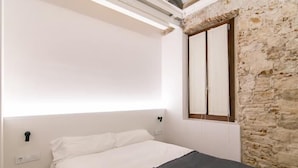 A bedroom with a bed, a wooden ladder, a stone wall, and a window with blinds. - Mid-term apartment 13 (Barcelona)