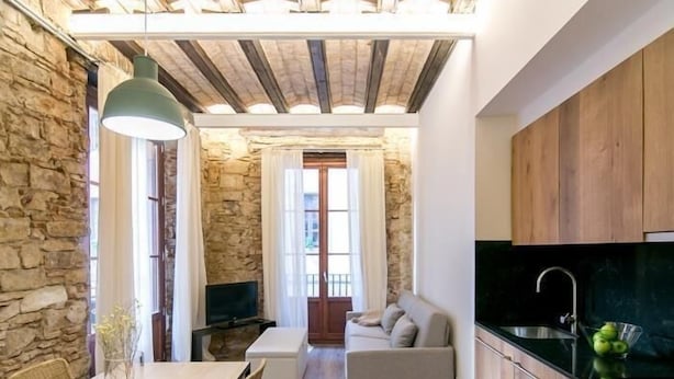 A modern kitchen with a black countertop and stainless steel sink, a dining area with a wooden table and chairs, and a living area with a sofa and a TV. - Mid-term apartment 13 (Barcelona)