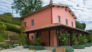 Una villa rosa con un porche cubierto, rodeada de vegetación y un camino de entrada de grava.