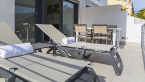 A sunlit outdoor seating area with lounge chairs and a table, a sliding glass door leading to a patio, and a view of the surrounding landscape. - Idaira II new spacious apartment with private garage and beach walking distance (Los Cristianos)