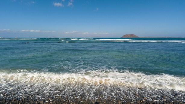 A beach with clear blue water, waves, and a distant mountain.