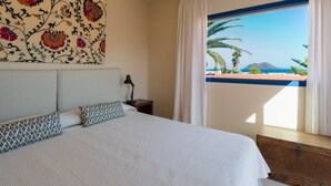 A bedroom with a large bed, a nightstand, a lamp, a window with curtains, and a colorful wall tapestry.