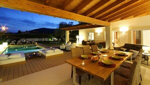 Ein Essbereich im Freien mit Holztisch und -stühlen, einem Pool und einer gemütlichen Sitzecke mit Kissen.